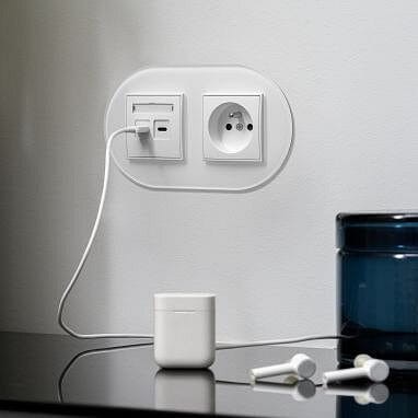 Ramka podwójna okrągła Karlik ICON w kolorze białym, zamontowana na ścianie, z widocznym gniazdem zasilającym oraz portem USB. W tle znajduje się szklany pojemnik i słuchawki. Kabel ładowarki podłączony do gniazda.