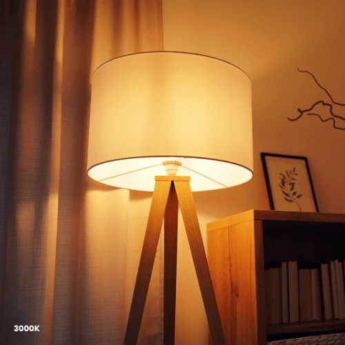 Żarówka LED filament E27 G45 4W w złotym kolorze, umieszczona w lampie stojącej z jasnym abażurem, emitująca ciepłe światło (3000K). Widoczny drewniany stelaż lampy i elementy dekoracyjne w tle.