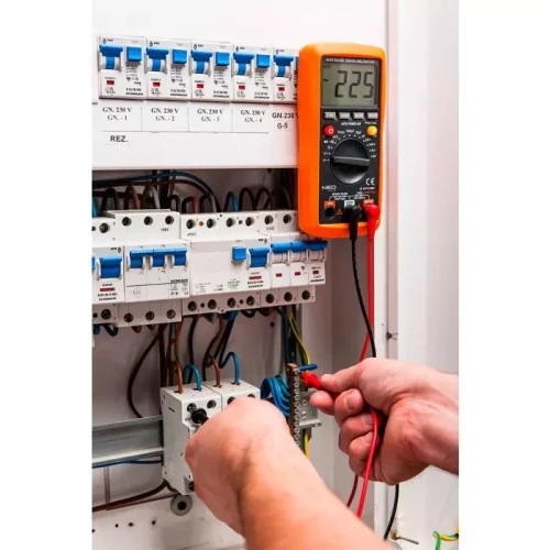 Miernik elektroniczny uniwersalny Neo 94-001 w kolorze pomarańczowym, z wyświetlaczem pokazującym wartość 225. Użytkownik trzyma przewody pomiarowe, zbliżając je do elementów instalacji elektrycznej w rozdzielnicy.