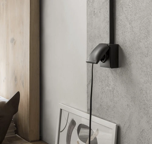 Gniazdo pojedyncze schuko w kolorze czarnym z klapką, zamontowane na ścianie. Produkt ma prosty, nowoczesny design, z widocznym kablem. Tło w odcieniach szarości z drewnianymi elementami i obrazem w ramie.
