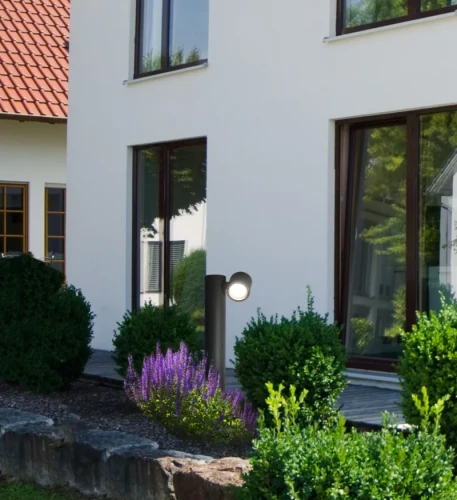 Lampa ogrodowa stojąca słupek PINO GU10 o wysokości 44 cm, w kolorze czarnym, umieszczona w ogrodzie obok krzewów i kwiatów, z widoczną głowicą oświetleniową skierowaną w górę. Tło to nowoczesny dom z dużymi oknami.