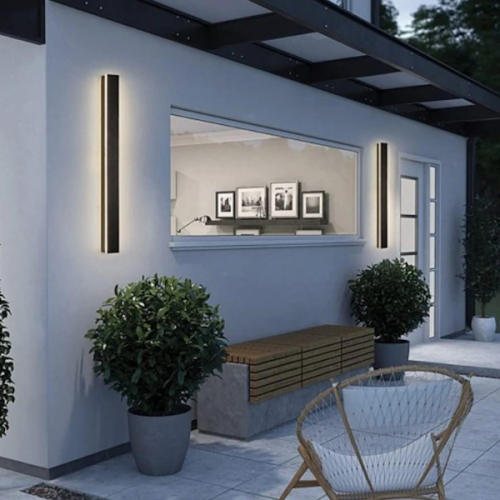 Oprawa elewacyjna LED LARA 45W, umieszczona na zewnętrznej ścianie budynku. Dwa prostokątne, czarne kinkiety z delikatnym podświetleniem, otoczone przez rośliny w donicach. W tle widoczna szklana witryna z ramkami na zdjęcia.