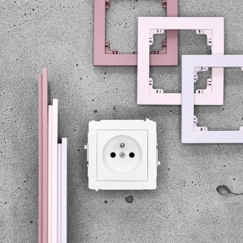 Pojedyncza ramka Karlik Deco w kolorze pudrowego różu, z kilkoma wariantami obramowania w odcieniach różu i fioletu, umieszczona obok białego gniazdka elektrycznego na szarym tle. Ramki mają prostokątny kształt i są montowane na ścianie.