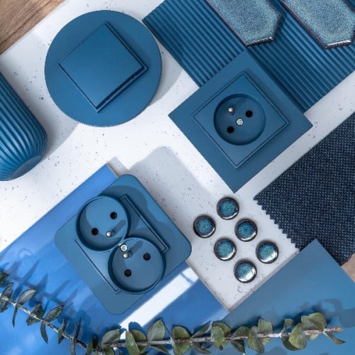 Łącznik pojedynczy Karlik MINI w kolorze navy blue, kwadratowy, z widoczną gniazdem elektrycznym. Produkt leży na jasnym tle, w otoczeniu innych elementów dekoracyjnych w odcieniach niebieskiego. Zauważalne detale to tekstura i wykończenie.