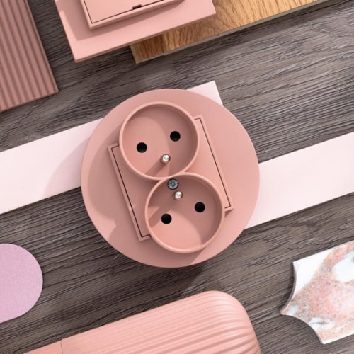 Gniazdo podwójne Karlik MINI w kolorze łososiowym, z uziemieniem. Okrągła obudowa z dwoma otworami na wtyczki. Widoczna metalowa śrubka. Produkt leży na drewnianym tle, otoczony innymi elementami dekoracyjnymi w pastelowych odcieniach.