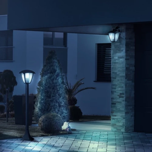 Lampa ogrodowa słupek IGMA o wysokości 86 cm, w kolorze czarnym, z okrągłą podstawą i kloszem w formie lampionu. Umieszczona w ogrodzie obok krzewów, tworzy przyjemne oświetlenie nocne. Tło stanowi nowoczesny budynek i kostka brukowa.